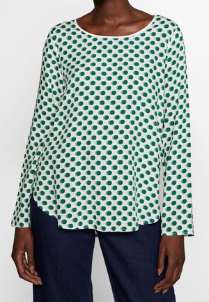 Femme portant un haut blanc à manches longues avec des pois verts et un pantalon bleu foncé, debout devant un fond clair uni.
