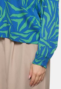 Niebieska bluzka z zielonym, abstrakcyjnym wzorem liści, z elastycznymi mankietami i luźnym krojem. Tkanina ma subtelną fakturę.
