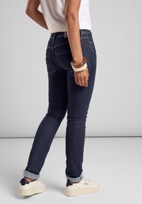 Mörkblå skinny jeans med stickningsdetaljer, uppvikta cuffs och en framträdande bakficka. Matchas med vita sneakers och en ljus topp.