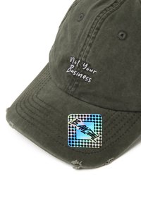 Cappellino da baseball in cotone verde oliva con testo ricamato "Not Your Business" e adesivo olografico con bordo a scacchi. Dettagli usurati.