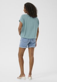 Luftig geschnittenes hellblaues Oberteil mit kurzen Ärmeln, kombiniert mit Denim-Shorts. Die Vorlage trägt weiße Blockabsatzsandalen, die einen strukturierten Stoff präsentieren.
