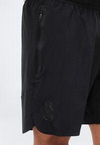 Μαύρο αθλητικό σορτς με πλαϊνή τσέπη με φερμουάρ και ανάγλυφο λογότυπο της Ρεάλ Μαδρίτης κοντά στο τελείωμα, φορεμένο στο πόδι ενός ατόμου πάνω σε απλό φόντο.