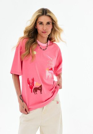 Kobieta o długich blond włosach, ubrana w różowy T-shirt z grafiką pustyni i zwierząt oraz jasne spodnie, pozująca na białym tle.