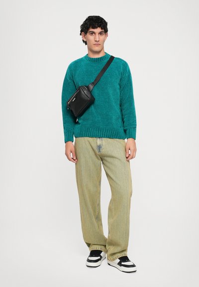 Young man stands wearing teal knit sweater, light yellow wide-leg jeans, black and white sneakers, and black crossbody bag against white background.