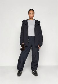 Ženska v črni smučarski jakni in hlačah, s sivo majico in črnimi smučarskimi čevlji, drži črne smučarske očala in stoji ob belem ozadju.