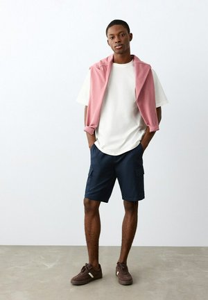 Mlad moški stoji z rokami v žepih, oblečen v belo majico, roza pulover, povešen čez ramena, mornarsko modre cargo kratke hlače in rjave superge.