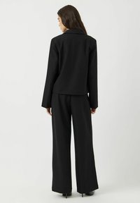 Černý přizpůsobený sak s dlouhými rukávy v kombinaci s volnými černými kalhotami. Hladká tkanina, strukturovaný design a minimalistický styl.