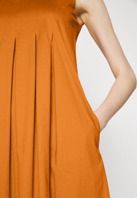 Robe orange sans manches avec des plis devant, montrant une main placée dans la poche latérale sur un fond neutre.
