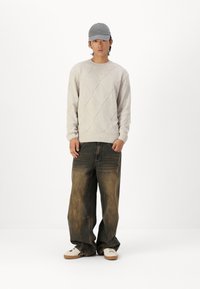 Light beige knitted sweater with diamond pattern, baggy faded blue jeans, and white sneakers with tan soles, paired with a gray cap.