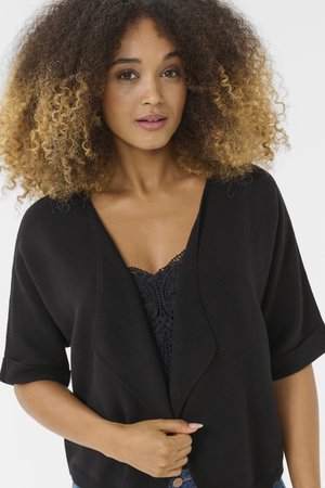 Kobieta z bujnymi kręconymi włosami ubrana w czarny kardigan na czarnej koronkowej bluzce, trzymająca kardigan zapięty na wysokości talii.
