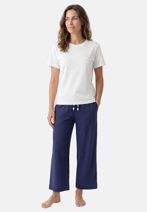 Vrouw staat op blote voeten, draagt een wit T-shirt met korte mouwen met een klein glimlachend gezicht en loszittende marineblauwe broek met trekkoord.