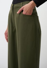 Pantaloni verde a gamba larga realizzati in tessuto resistente. Presentano tasche laterali e una vita pulita. Texture liscia con dettagli di cucitura sottili.