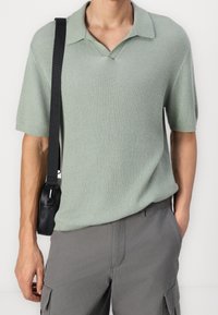 Man som bär en ljusgrön kortärmad stickad polo skjorta och grå cargo shorts, med ett svart axelrem som syns på vänster sida.