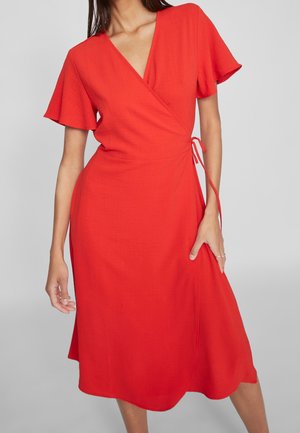Kobieta ubrana w czerwoną sukienkę o długości do kolan, z krótkim rękawem i wiązaniem po boku, stojąca na tle jednolitego, jasnoszarego tła.