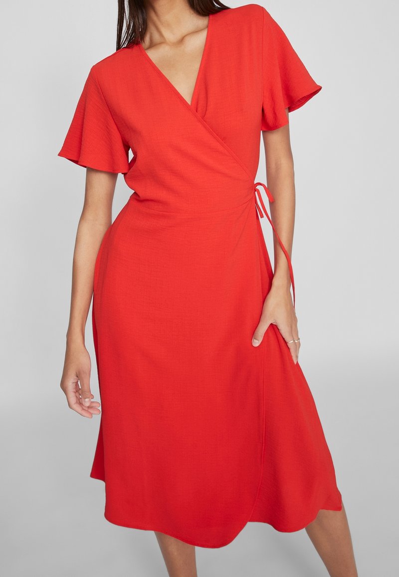 Femme portant une robe portefeuille rouge à manches courtes arrivant au genou avec un nœud sur le côté, debout devant un fond gris clair uni.
