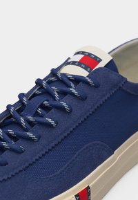 Μπλε ναυτικό αθλητικό παπούτσι με κορδόνια άσπρης ρίγας, επάνω μέρος από υφή σουέντ και ύφασμα, λευκή σόλα και λογότυπο Tommy Jeans σε κόκκινο, άσπρο και μπλε στη γλώσσα και στο πλάι.