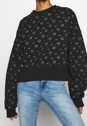 Svart sweatshirt med gyllene cirkulära mönster och Nike-logotyper, med en croppad design och ribbade manschetter. Kombineras med ljusblåjeans.