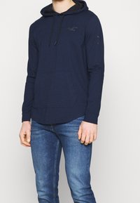 Marinblå hoodie med känguruficka, långa ärmar och en subtil logo. Tillverkad av mjukt tyg med en slät struktur.