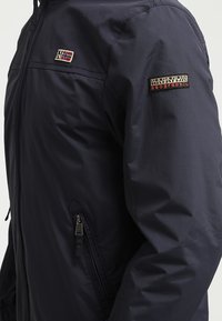 Marinblå vattentät jacka med dragkedja och sidoficka. Vänstra axeln har en liten norsk flagga-lapp och högra axeln har en logotyp-lapp.