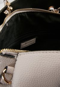 Avoin vaaleanbeige tekstiilitasku, jossa on kultaset vetoketju ja ketjukahva, paljastaen sileän mustan kankaan sisäpuolen ja sisäisen vetoketjullisen taskun.