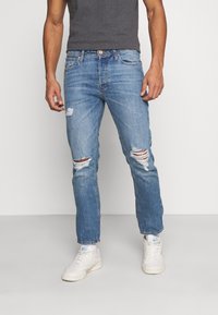 Jeans de ganga azuis com um corte descontraído, apresentando detalhes desgastados e rasgos nos joelhos, usados com ténis brancos.