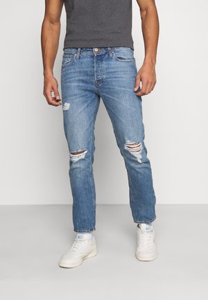 Jeans de ganga azuis com um corte descontraído, apresentando detalhes desgastados e rasgos nos joelhos, usados com ténis brancos.