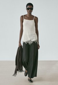 Schlanke Person mit Sonnenbrille, trägt weißes Spitzentop und weite schwarze Hose, hält einen dunklen, mit Fransen versehenen Schal, geht auf hellem Boden vor hellem Wandhintergrund.