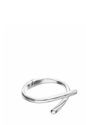 Bracciale color argento, aperto, con un design moderno e curvo, posato su una superficie bianca, che mostra la sua finitura metallica liscia.
