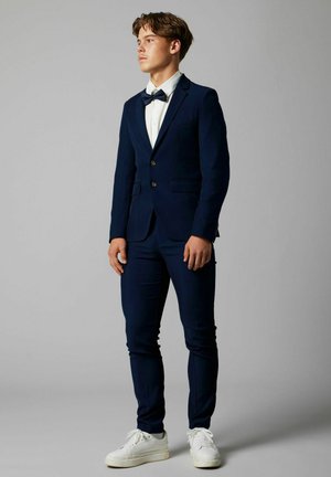 Completo blu con giacca a un solo petto, due bottoni e rever a scollatura. Abbinato a una camicia bianca, cravatta a farfalla nera e sneaker bianche.