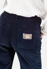 Marineblauwe corduroy broeken met een patchzak aan de achterkant met een bloemenontwerp en een "cache coeur" label. Getextureerde ribbelstof.