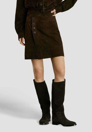 Femme portant une jupe boutonnée en daim marron foncé, un haut à manches longues assorti et des bottes en daim montant jusqu'aux genoux, debout avec une main dans la poche.