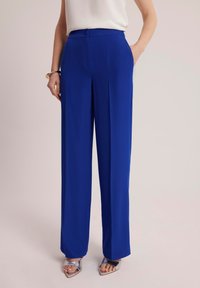 Kobaltowe, szerokie spodnie z gładkiego, lekkiego materiału, z plisowanym przodem i zapięciem na guziki w talii. Widać srebrne buty akcentowe.