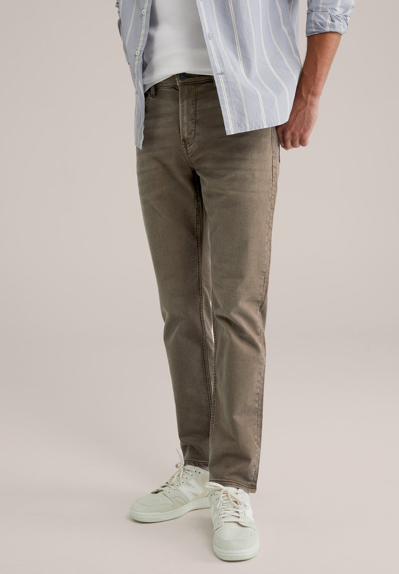 Gai pelēks svītrains krekls pār piemērotu baltu t-kreklu, kombinēts ar olīvzaļām džinsām un baltām sporta kurpēm. Gluda auduma, ikdienas dizains.