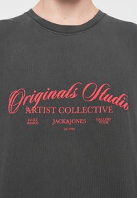 Tumši pelēks kokvilnas T-krekls ar izteiksmīgu sarkanu grafiku tekstā, kas ietver "Originals Studio" un "Artist Collective", ar apaļu dekoltē.