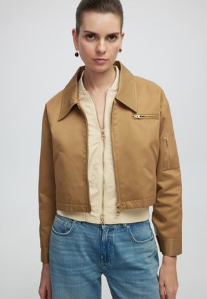 Korte bruine jas met een brede kraag en ritszakken, gelaagd over een crèmekleurige zip-up top, gecombineerd met lichtblauwe jeans.