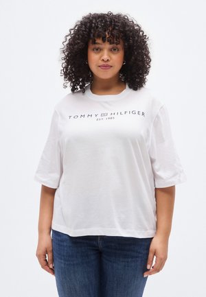 Donna con capelli ricci che indossa una t-shirt bianca Tommy Hilfiger e jeans blu, in piedi contro uno sfondo bianco uniforme.