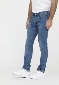 Jean en denim bleu avec une coupe slim, cinq poches et une fermeture éclair. Le matériau a une texture douce, avec des décolorations et des détails de couture subtils.