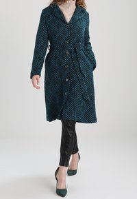 Cappotto lungo color teal con un motivo a rombi, colletto ampio, bottoni e cintura da annodare. Indossato sopra pantaloni neri aderenti e tacchi verdi.