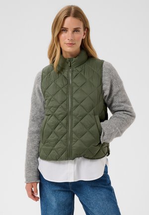 Donna che indossa un gilet verde trapuntato sopra un maglione grigio e una camicia bianca, posando con una mano in tasca davanti a uno sfondo semplice.