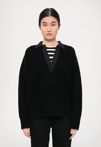 Schwarzer gestrickter Pullover mit V-Ausschnitt und kontrastierendem schwarzen Kragen, getragen über einem schwarz-weiß gestreiften Hemd. Die Passform ist locker und entspannt.