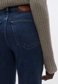 Sötétkék farmernadrág sima textúrával, két hátsó zsebből és barna bőr övsáv címkével. Látható kötött pulóver részlet.