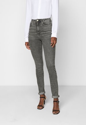 Person iført opkørte grå skinny jeans, hvid langærmet skjorte med knapper og sorte højhælede sandaler, stående på ensfarvet baggrund.