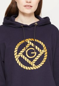 Sudadera azul marino con un patrón de cuerdas doradas que presenta un logo central. Cuerdas con herrajes dorados; material de algodón suave.