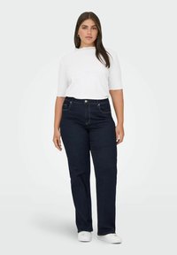 Tumši zili džinsi ar taisnu kāju dizainu, kuriem ir piecas kabatas un kontrastējoša šuvuma, savienoti ar baltu, ribotu īsām piedurknēm topu.