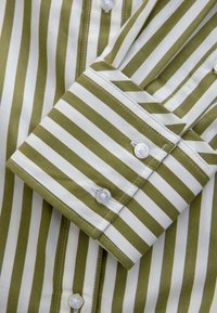 Manche retroussée d'une chemise à rayures verticales vertes et blanches, boutonnée avec des boutons blancs et des coutures visibles.