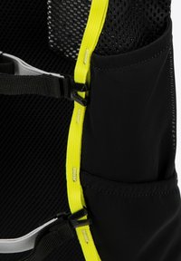 Zaino nero con pannello in rete, accenti gialli e vari cinturini. Presenta una tasca e fibbie in plastica per la regolazione. Tessuto strutturato.