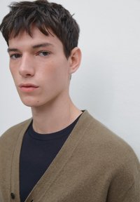 Light brown cardigan with v-neck design, button closure, worn over a dark navy shirt. Smooth texture, minimalistic style against a light background.