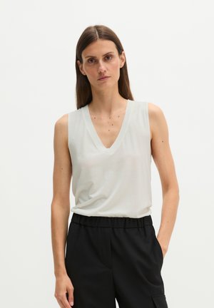 Femme aux cheveux bruns raides portant un haut blanc sans manches à col en V et un pantalon noir, une main dans la poche sur fond uni.