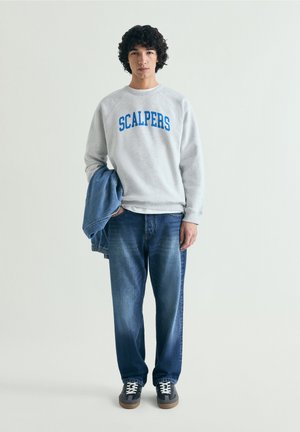 Jeune homme portant un sweat-shirt gris avec le texte "SCALPERS", un jean bleu et des baskets noires, tenant une veste en jean dans une main.