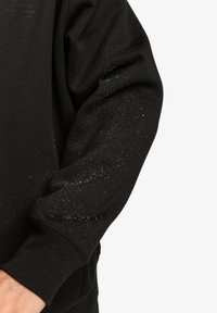 Felpa nera con maniche lunghe, realizzata in misto cotone, con un motivo testurizzato di piccole macchie sulle maniche. Visibile il marchio Elements.